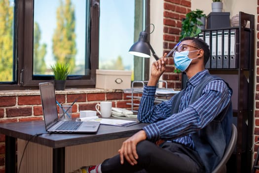 African american manager seated in brick wall office looking pensive and tapping his forehead with a pen. Businessman with face mask, gazing outside the window and thinking deeply on company projects.
