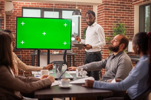 Digital monitor displaying blank chromakey mockup template as black businessman stands nearby doing presentation for team. African american manager speaking to coworkers, next to device with greenscreen.