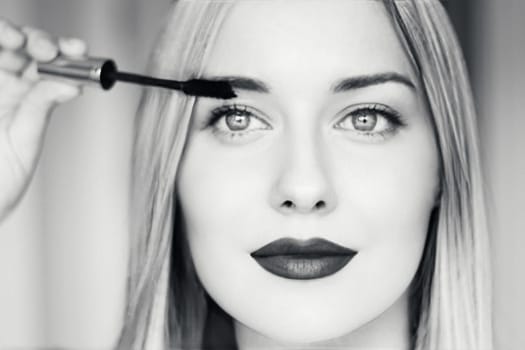 Close-up of a beautiful blonde woman applying mascara, with dark lipstick, bright eyes, and long straight hair. Black and white portrait for beauty, makeup, fashion, or cosmetic themes
