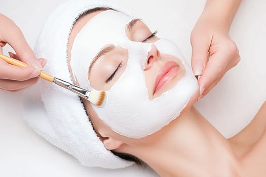 An elderly woman lies comfortably on her back in a calming spa environment as a beautician applies a soothing white mask to her face with a brush. Soft light illuminates the serene atmosphere, enhancing relaxation.