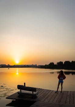 A woman standing on a wooden dock, gazing at a serene sunset over a calm lake with reflections and a tranquil atmosphere.