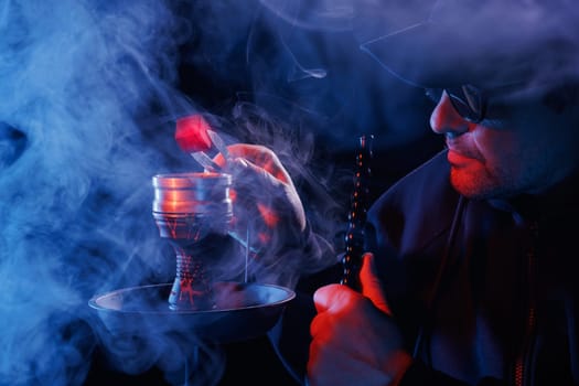 Man smoking hookah, adjusting hot charcoal with tongs in a smoky, moody atmosphere, illuminated by red and blue lighting,