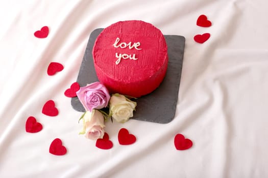 Romantic red cake with love message surrounded by roses and hearts on fabric.