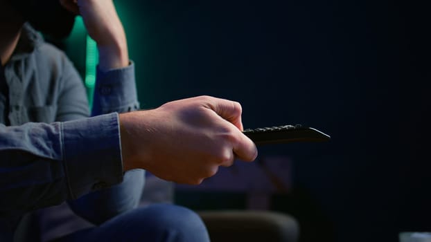 Close up of man on couch using remote control to browse though movies available on streaming service. Focus on remote control used by homeowner in apartment switching channels on TV, camera B
