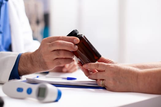 Closeup of white person in lab coat giving medicine to pensioner patient after medical exams. Selective focus of caucasian people holding pill bottle on table with thermometer and clipboard nearby.
