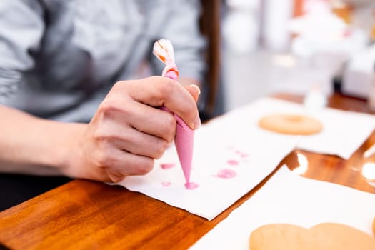 Adult female decorating cookies with pink icing using a piping bag in a kitchen setting.