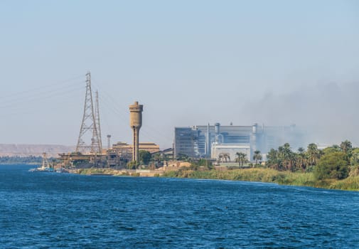 A view of the Edfu steel factory, including industrial buildings, smokestacks, and power lines, seen from the Nile River