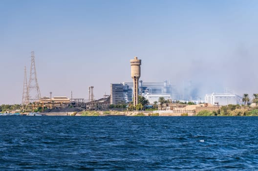A view of the Edfu steel factory, including industrial buildings, smokestacks, and power lines, seen from the Nile River