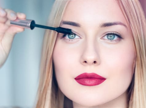 Woman applying mascara with confidence, defined brows and red lipstick. Blonde beauty focused on her eye makeup, cosmetics look