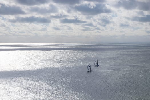 Aerial view of two sailboats plowing together in calm sea. Nearby, kite surfers soar with colorful sails, carving waves and leaping into the air