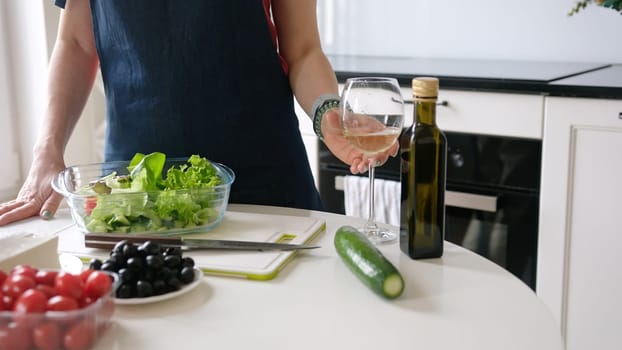 Young Woman Enjoys White Wine At Home In The Kitchen While Preparing Greek Salad, Embracing Home Cooking