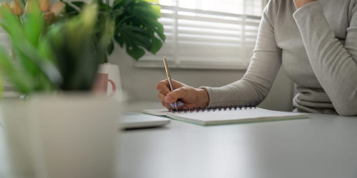 Focusing intently, an individual writes in a notebook at their desk, surrounded by plants and light, illustrating a serene work-from-home atmosphere.