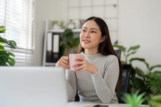 In a cozy home office, a thoughtful young woman enjoys a cup of tea, reflecting on her upcoming workday. The setting is filled with greenery and light.