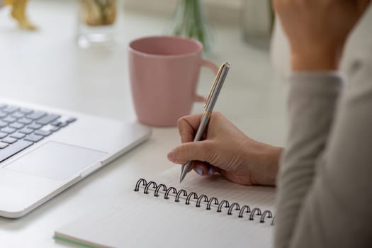 Focusing intently, a woman writes in her notebook next to a laptop, demonstrating the balance of writing and digital work while sipping coffee.