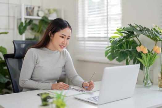 A young woman actively participates in a virtual meeting, jotting down key points in her notebook amidst a bright, plant-filled workspace.