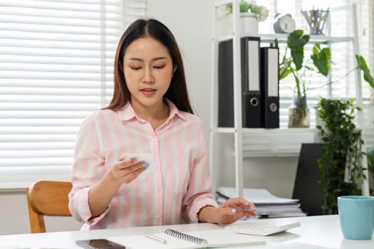 A young woman engages in focused work, reviewing information on her smartphone while surrounded by her home office setup.