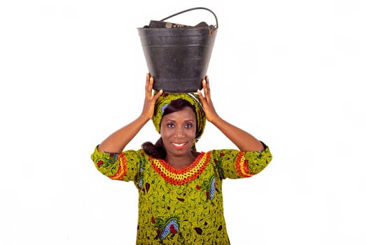 beautiful african adult woman dressed in green loincloth, balancing a black bucket on her head with her hands, smiling