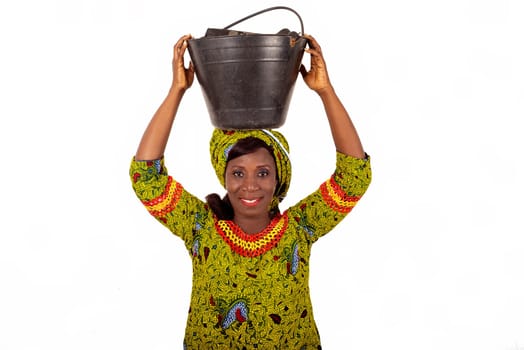 beautiful african adult woman dressed in green loincloth, balancing a black bucket on her head with her hands, smiling