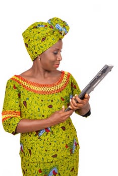beautiful african adult woman dressed in loincloth and green scarf is standing, holding a laptop computer in hand while being smiling