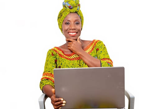 Beautiful African adult woman wearing a loincloth and a green scarf on her head, sits on a chair with her computerist smiling to put her hand on her chin