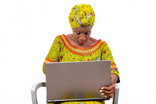 Beautiful African adult woman wearing a loincloth and a green scarf on her head is sitting on a chair manipulating her computer
