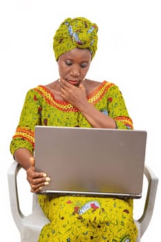 Beautiful African adult woman wearing a loincloth and a green scarf on her head, sits on a chair with her computer thinking about putting her hand on her chin