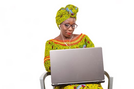 beautiful african adult woman dressed in loincloth and green scarf with glasses, is sitting on a chair with her computer on her feet, and takes pleasure in communicating with headphones smiling