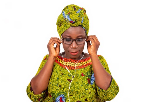 beautiful african adult woman dressed in loincloth and green scarf with glasses, is sitting on a chair, and enjoys listening to music with headphones while smiling