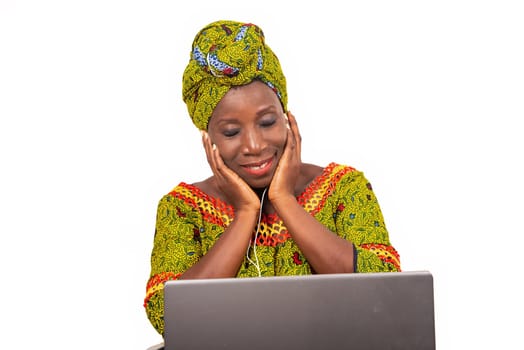 beautiful african adult woman dressed in loincloth and green scarf, enjoys listening to music with headphones, in her laptop while smiling with her eyes closed.