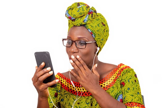 beautiful african adult woman dressed in loincloth and green scarf with glasses, takes pleasure listening to music with headphones, in her cellphone.