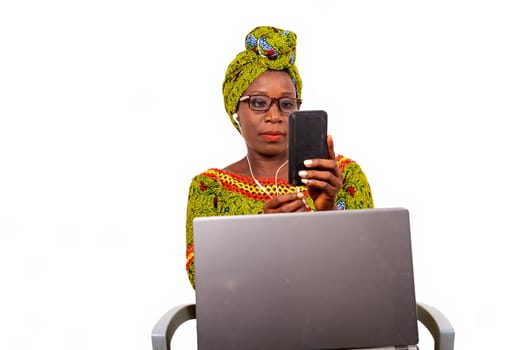 beautiful african adult woman dressed in loincloth and green scarf with glasses, is sitting on a chair with her computer on her feet, and enjoys communicating with headphones, in her cellphone