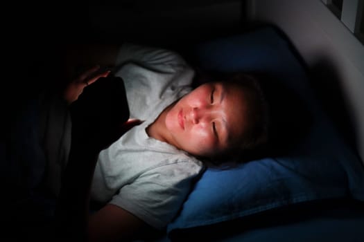 Asian woman lying in bed using smartphone at night, illuminated by screen light