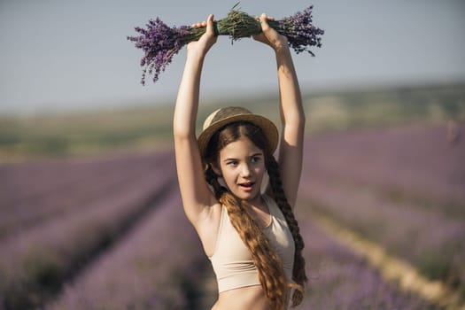 girl is holding a bunch of lavender purple flowers in her hands and wearing a straw hat. She is smiling and she is enjoying herself. The scene is set in a field of lavender, which adds to the peaceful