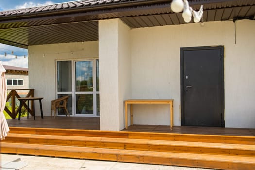 Wooden steps lead to porch with black door and wicker chair. Simple architectural structure and functional outdoor space usage
