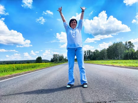 Middle-aged blogger celebrates her journey by stretching arms wide on a picturesque country road under a bright blue sky.