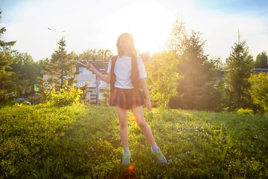 A girl wears a white shirt and a plaid skirt, holding a stick against the backdrop of a vibrant sunset and lush greenery.