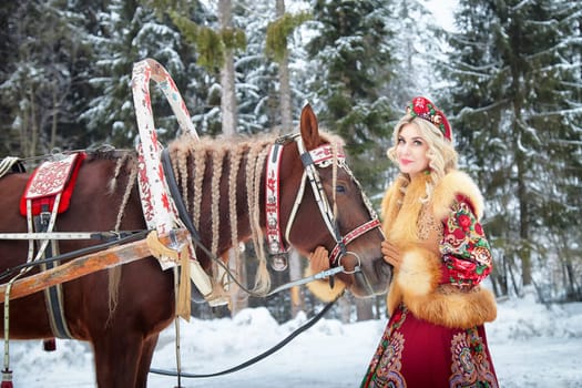 A beautiful Russian girl in vibrant attire poses with a horse, celebrating Maslenitsa in a winter setting.