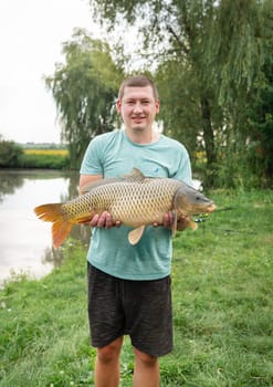 A fisherman proudly holding a large carp near a scenic lake, surrounded by lush greenery and a calm outdoor setting.