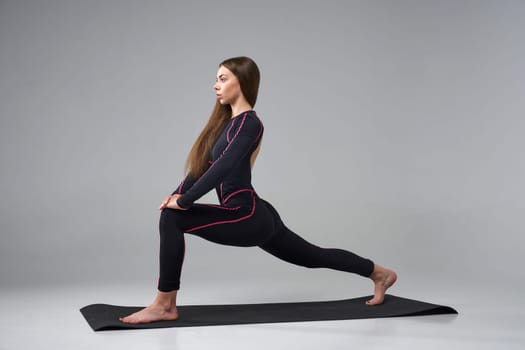 Slim woman in black bodysuit exercising on gym mat. Side view of calm Caucasian girl with long hair, practicing yoga, isolated on grey studio background. Concept of yoga, sport, healthy lifestyle.