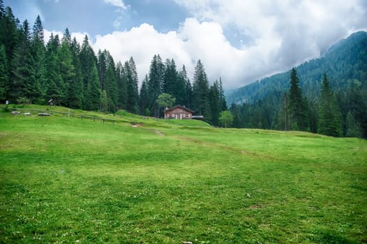 Lush green meadow with a wooden chalet in the distance, surrounded by the italian dolomites, Trentino Alto Adige