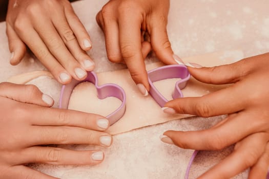 Four hands use a heart-shaped cookie cutter to cut out shapes from dough.
