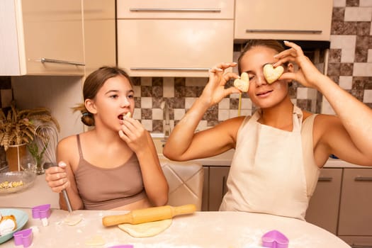 Two women in a kitchen, one holding dough over her eyes, the other biting into a piece. They are smiling and having fun.