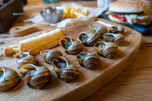 Delicious escargot and sliced bread on wooden board with burger in background.