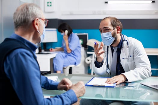 Male physician sits with pensioner patient, discussing health concerns and reviewing notes on clipboard in medical office. Young medic specialist describing healthcare treatment plans for senior man.