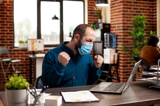 Male entrepreneur clenches fists in joy after presenting winning marketing strategy at office desk. Happy businessman with face mask sitting at table and celebrating successful company project deal.