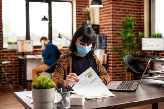 Businesswoman seated at tidy desk with laptop, examining detailed financial documents in brick wall room. Female analyst wearing face mask and reviewing startup company research paperwork on table.