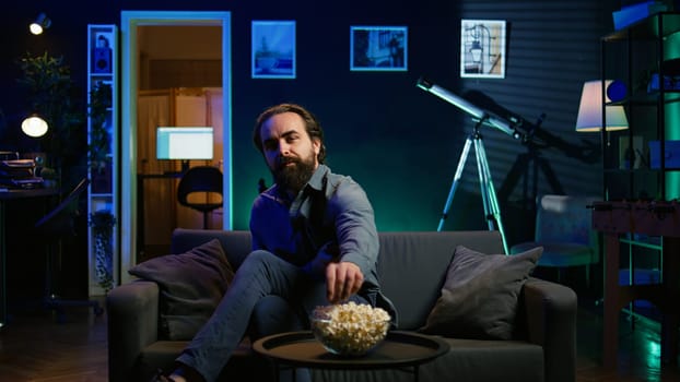 Man taking seat on sofa to watch television, getting entertained in relaxed home environment. Person arriving in apartment sitting down on couch to enjoy movies, turning on TV, eats popcorn, camera B