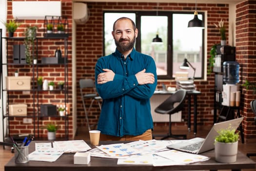 Portrait of young caucasian guy with arms crossed standing near table having financial documents in brick wall room. Bearded man in business casual clothing is looking at camera in startup office.
