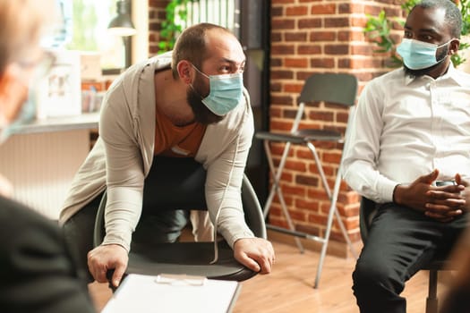 Young man in face mask expresses anger and despair about alcohol addiction amidst pandemic. Caucasian guy shouting during psychotherapy group session, having nervous breakdown in brick wall room.