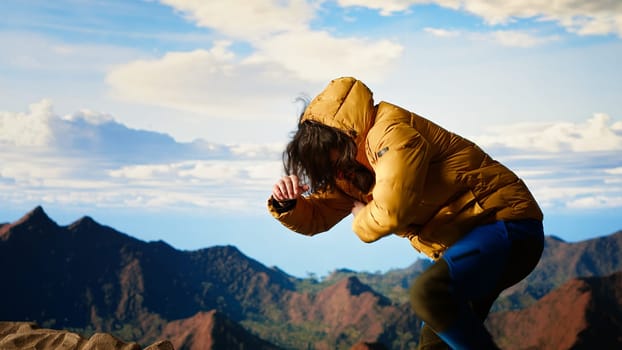 Clumsy traveler loses balance on a steep mountain peak as powerful winds knock him off course, falling and being unequipped in expedition. Unprepared hiker for the cold weather. Camera B.